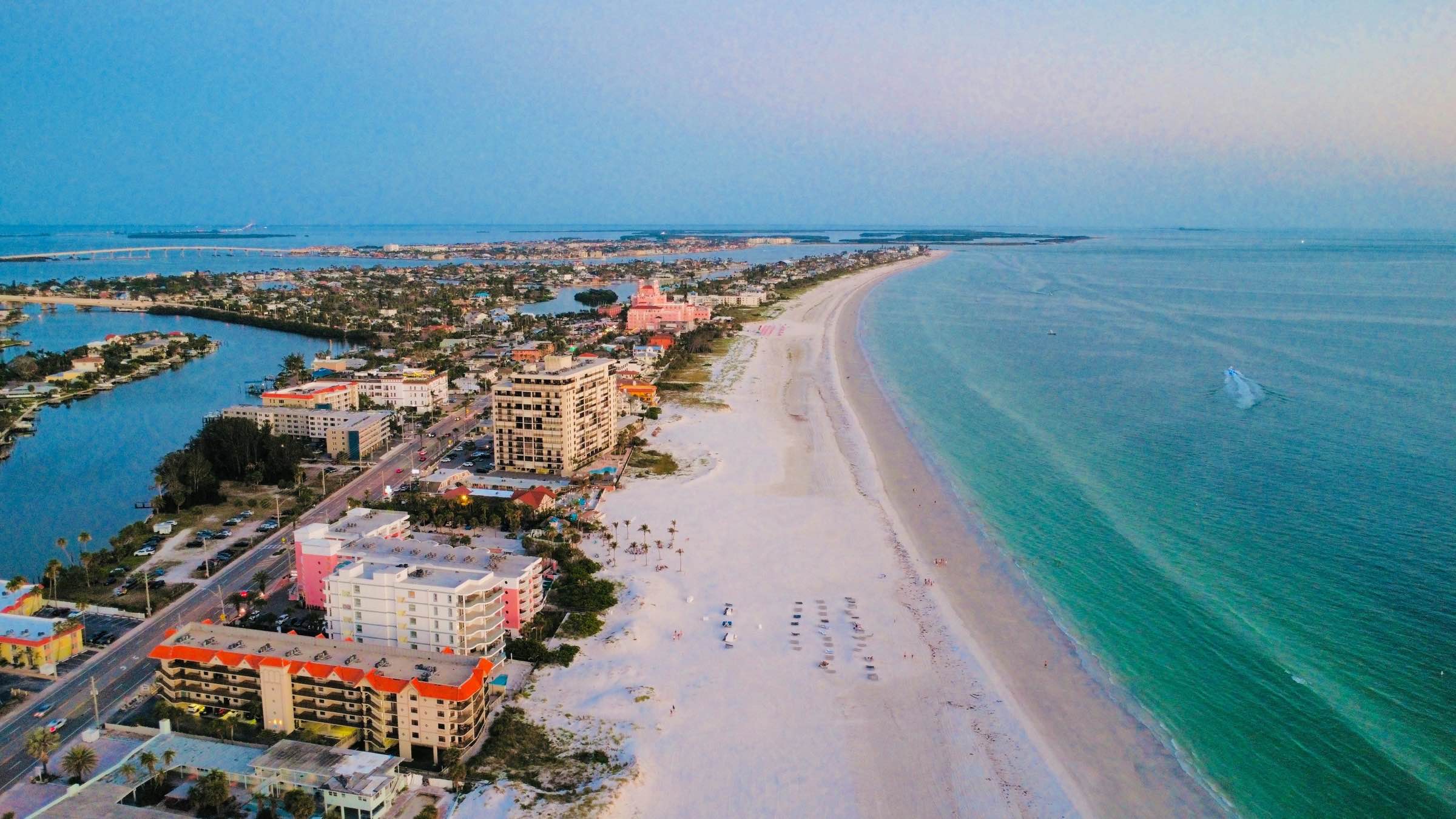 Aerial view along St. Pete Beach at dusk. Low-rise and mid-rise condominium buildings line Gulf Boulevard on the left, a wide white sand beach with rows of beach chairs runs through the center, and the turquoise Gulf of America fills the right side. The pink Don CeSar hotel stands mid-frame, with Boca Ciega Bay and the barrier island stretching to the horizon.