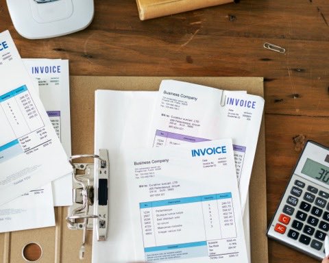 Invoices and receipts spread across a desk beside a binder, a calculator and a phone, with a person reviewing the paperwork