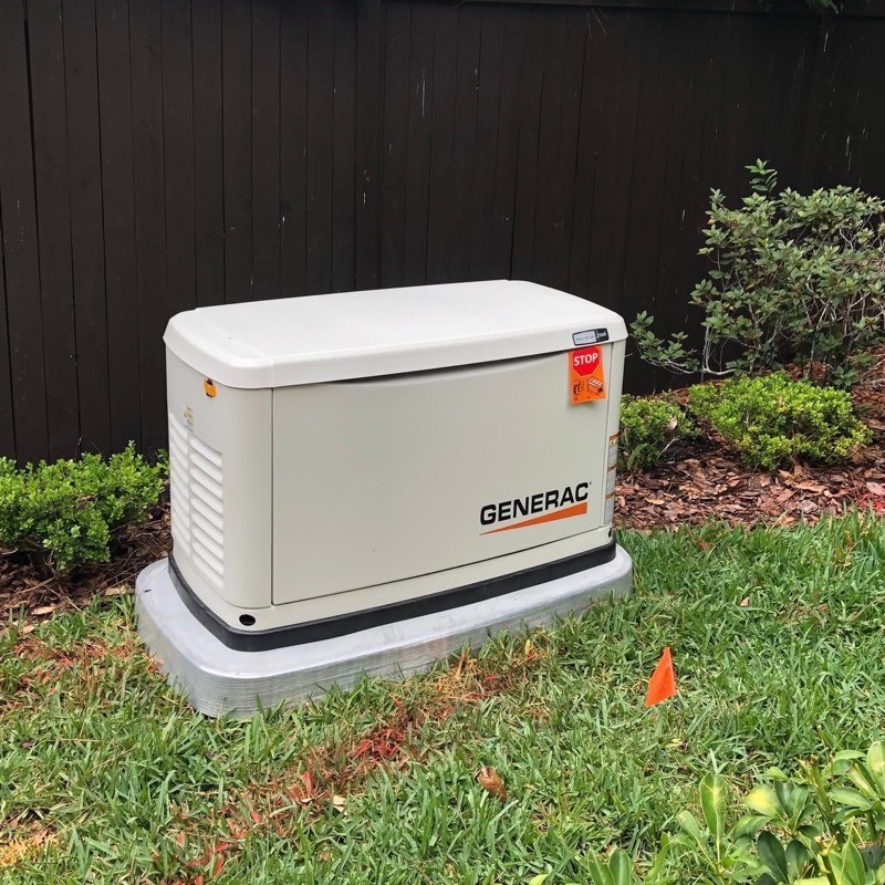 A Generac standby generator installed on a concrete pad beside a dark wood fence, with utility marking flags in the grass
